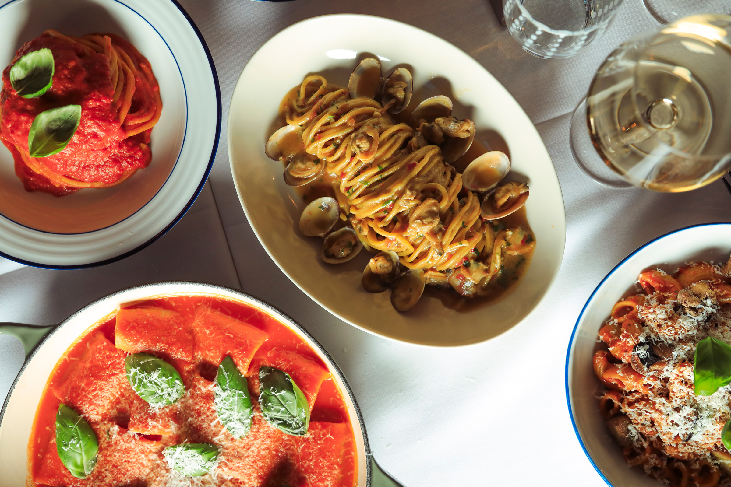 A guest lifting a forkful from an oval dish of ridged pasta in tomato sauce, finished with basil and grated cheese, beside a glass of white wine on a crisp white tablecloth.