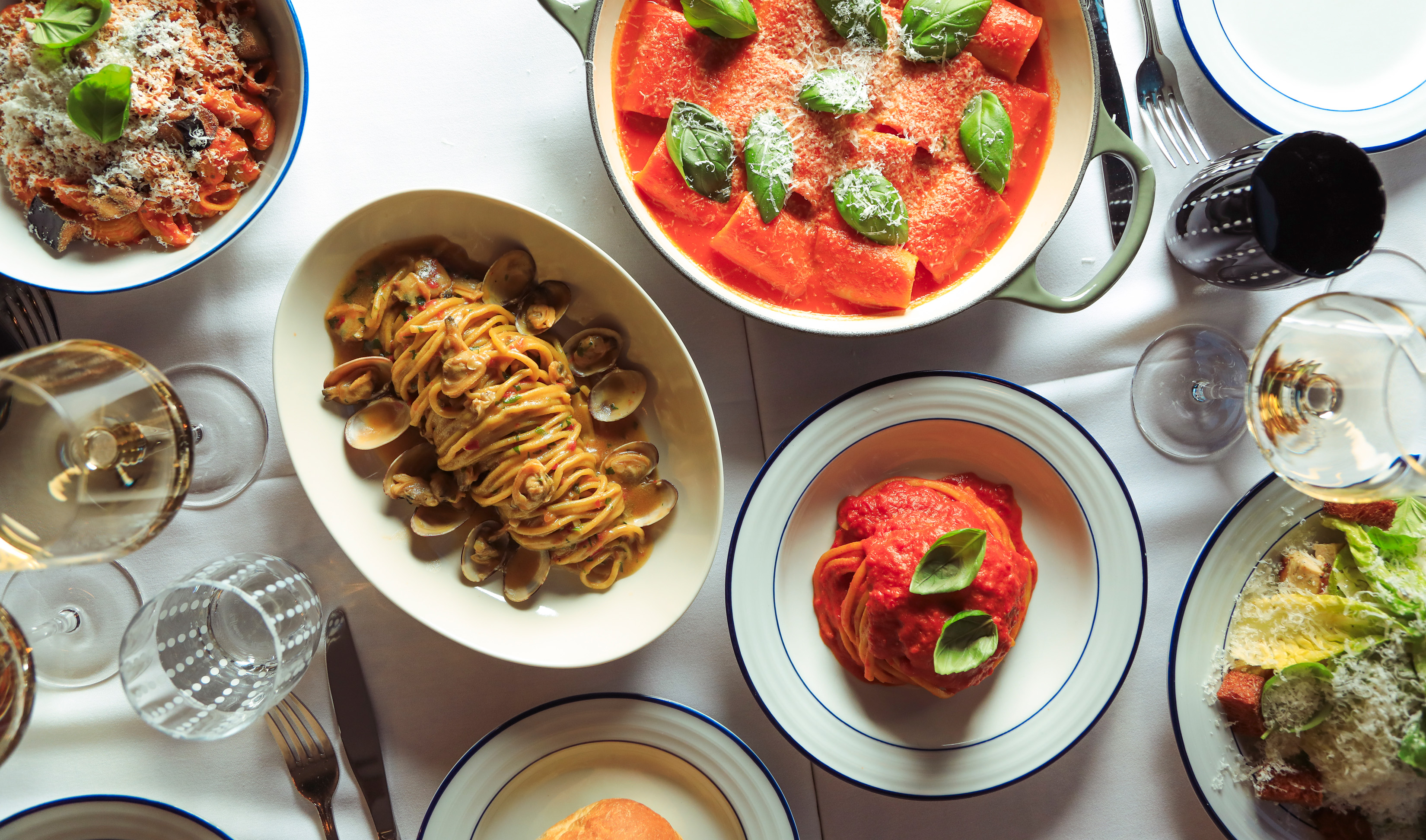 A sunlit table laid with Ribalta pasta dishes seen from above: spaghetti alle vongole in a shallow bowl, a baked tomato pasta scattered with basil and grated cheese, and a glass of white wine.