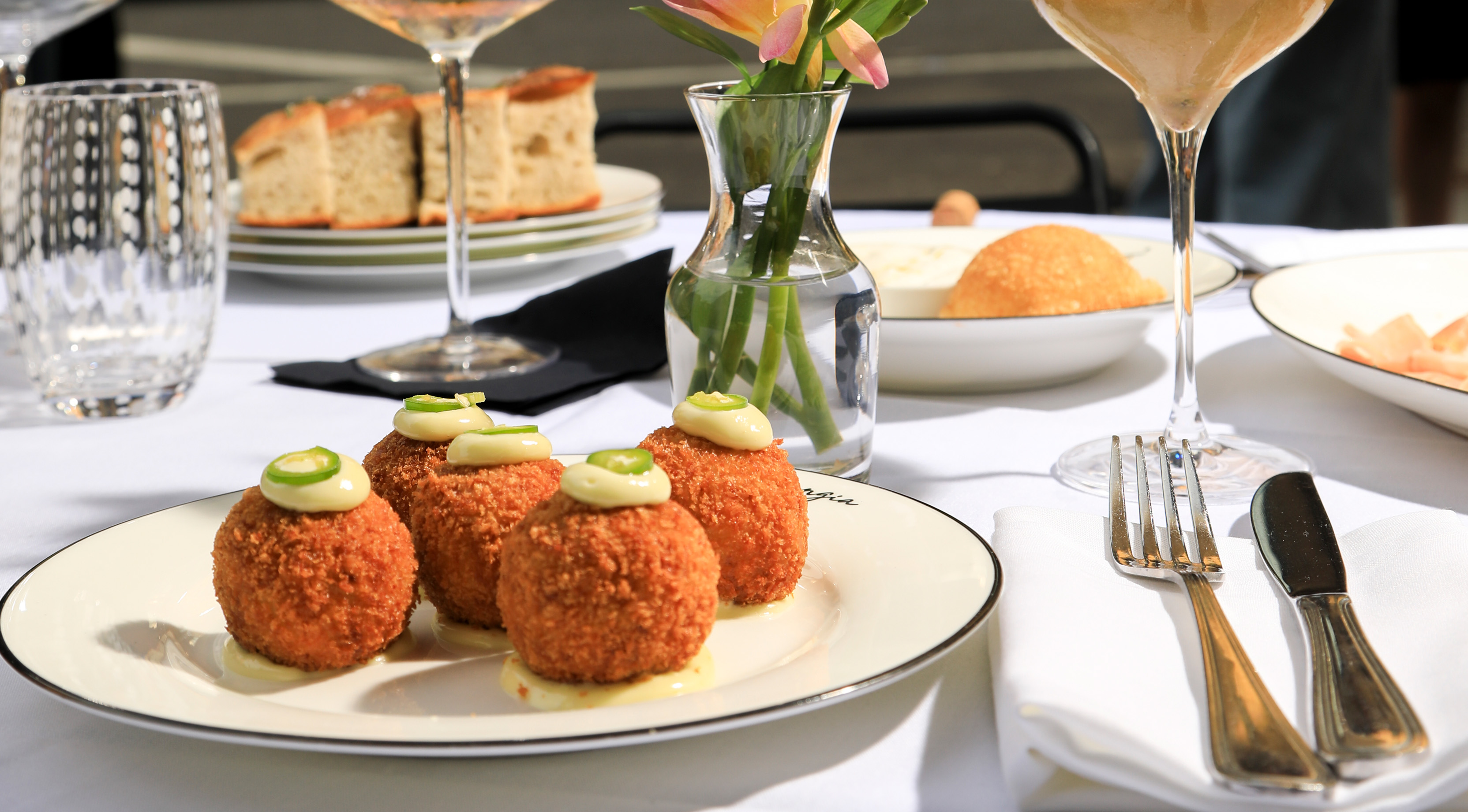 A guest lifting a forkful from an oval dish of ridged pasta in tomato sauce, finished with basil and grated cheese, beside a glass of white wine on a crisp white tablecloth.